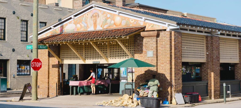 Charleston City Market building with brick structure, open-air vendor stalls, and people setting up goods under the covered walkway.